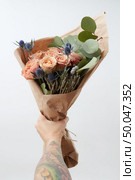 Summer wonderful fresh roses, eryngium and green leaves as a greeting bunch in a female’s hand with tattoo on a gray background. Place for text. Gift for Mother’s Day.. Woman’s hand with tattoo is holding beautiful colored living coral rores bouquet. Greeting card for Mother’s or Woman’s Day. Copy space. Стоковое фото, агентство Ingram Publishing / Фотобанк Лори