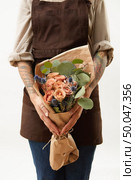 Beautiful flowers coral living roses and eryngium as a congratulate bouquet in girl’s hand with tattoo, place for text. Concept of Mother’s or Woman’s Day.. Woman in a brown apron is holding summer wonderful fresh roses, eryngium and green leaves as a greeting bunch on a light background. Greeting card. Copy space. Стоковое фото, агентство Ingram Publishing / Фотобанк Лори