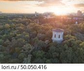A city in the distance and a green park with a tower and people on the roof against the sky with the sunset. Aerial view from the drone. Aerial view of the drone on the green park with a round tower and the city in the distance at sunset. Стоковое фото, агентство Ingram Publishing / Фотобанк Лори