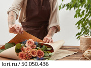 Florist woman in a brown apron is wrapping fresh rose bouquet in a decorative paper on a wooden table on alight background. Process step by step. Concept of floral shop.. Gardener girl make bouquet for a holiday from living coral roses and branches of green leaf on a wooden table on a light background. Mother’s Day. Стоковое фото, агентство Ingram Publishing / Фотобанк Лори