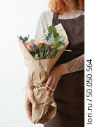 Florist woman’s hands with tattoo hold flower roses bouquet for congratulation on a gray background. Concept of Mother’s or Woman’s Day. Place for text.. Female in a brown apron with red hair hold nice bouquet from living coral colored roses and eryngium on a light. Стоковое фото, агентство Ingram Publishing / Фотобанк Лори