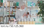 Mature woman chooses a medicinal cough syrup in a pharmacy. Elderly European woman buys syrup for sore throat and cough for her grandchildren during cold and flu season. Стоковое видео, видеограф Яков Филимонов / Фотобанк Лори