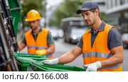 Two male waste management workers in bright safety vests are collecting garbage bags from green bins on a city street, showcasing teamwork and environmental responsibility. Стоковое AI-изображение, AI-художник Ирина Ручушкина / Фотобанк Лори