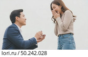 A young Asian man kneels with a ring, proposing to a surprised young Asian woman. She covers her mouth in joy, set against a neutral background. Стоковое AI-видео, аниматор Ольга Невская / Фотобанк Лори