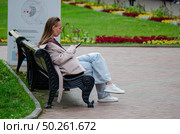 Девушка 30 лет с длинными русыми волосами, черными очками на лбу сидит на скамейке в белом плаще или пальто, голубых джинсах и белых кроссовках и смотрит что то в телефоне. Вокруг парк музей заповедник Коломенское, яркие цветочные клумбы и инсталляция с информацией. Редакционное фото, фотограф Светлана Русских / Фотобанк Лори