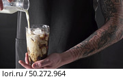 Cooking summer drink. A man with a tattoo pours milk into a glass with coffee and ice around a dark background. 4K UHD video, 240fps, 2160p.. A male barista with a tattoo pours fresh milk into a glass with ice and coffee around a dark background. Cooking Frappe. 4K UHD video, 2160p. Стоковое фото, агентство Ingram Publishing / Фотобанк Лори