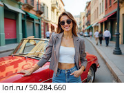 Молодая женщина в солнечных очках стоит возле красной машины на улице города. Стоковое AI-изображение, AI-художник Наталия Ануфриева / Фотобанк Лори