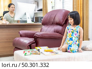 Asian girl child reading interactive book in living room at home as home schooling while her mom working at home because city lockdown because of covid-19 pandemic. Home Scholling concept. Стоковое фото, агентство Ingram Publishing / Фотобанк Лори