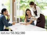 Asian waitress wear facemask and face shield using tablet to show restaurant electronic menu to reduce touching and recommend to customer. New normal technology and lifestyle food and dining concept. Стоковое фото, агентство Ingram Publishing / Фотобанк Лори