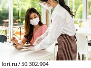 Asian waitress wear facemask and face shield using tablet to show restaurant electronic menu and recommend to customer. New normal technology and lifestyle food and dining concept. Стоковое фото, агентство Ingram Publishing / Фотобанк Лори