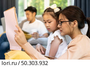 Asian girl reading tale story book with her mom in living room with dad work from home while city lockdown from coronavirus covid-19 pandemic. Domestic life family and work at home concept. Стоковое фото, агентство Ingram Publishing / Фотобанк Лори