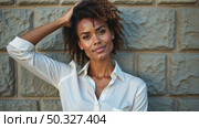 A black woman in a white blouse standing confidently with her arm raised against a textured stone wall, soft lighting enhancing. Стоковое AI-видео, аниматор Sergey Grishin / Фотобанк Лори