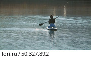A woman floating on a paddleboard on a calm body of water reflecting the sun's glare. She is wearing a black top and jeans. Back view. Riding on sup board. Стоковое видео, видеограф Воловодов Владимир / Фотобанк Лори