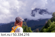 Young woman hiker in cap with orange backpack gazes at majestic mountains shrouded in clouds, surrounded by lush greenery, capturing the essence of adventure and exploration in nature. Стоковое фото, фотограф Елена Гоосен / Фотобанк Лори