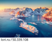 Aerial view of snowy islands with rorbu, sea, rocks in water and mountains at sunrise in winter. Beautiful landscape with village, road, bridge. Top view from drone. Hamnoy, Lofoten islands, Norway. Стоковое фото, агентство Ingram Publishing / Фотобанк Лори