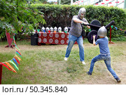Father and child wearing helmets engage in playful medieval sword fight with foam swords and shields on summer day. Стоковое фото, фотограф Алла Чесноковва / Фотобанк Лори