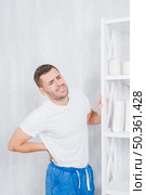 young man standing against concrete wall touching his back grimacing face with pain. Стоковое фото, агентство Ingram Publishing / Фотобанк Лори