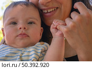 Молодая женщина держит на руках четырёхмесячную девочку в полосатой футболке на фоне густой зелёной изгороди в солнечном парке. Стоковое фото, фотограф Владислав Войтов / Фотобанк Лори