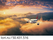 Airplane is flying above the clouds at sunset in summer. Landscape with passenger airplane, mountains, orange sky. Aircraft is taking off. Business travel. Commercial plane. Aerial view. Transport . Стоковое фото, агентство Ingram Publishing / Фотобанк Лори