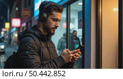 Hispanic young attractive man stands in dark street in front of shop, changes songs and tracks on smartphone, listens to music in wireless headphones. Hipster wearing casual dress. Стоковое AI-видео, аниматор Sergey Grishin / Фотобанк Лори