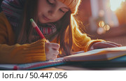 A girl in a knit hat and scarf focuses on writing in her notebook, a warm glow surrounding her. Стоковое AI-изображение, AI-художник Данила Большаков / Фотобанк Лори