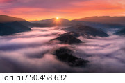 Aerial view of mountains in low clouds at sunrise in autumn. Top view of mountain hills in fog in fall at dawn. Beautiful landscape with ridges, forest, sun, colorful orange sky with pink clouds. Стоковое фото, агентство Ingram Publishing / Фотобанк Лори
