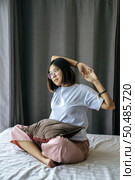 A woman in a white shirt sitting on the bed and raising both arms. Стоковое фото, агентство Ingram Publishing / Фотобанк Лори