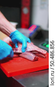 Professional butcher cutting lamb shank on red board, wearing blue gloves and handling raw meat with precision during butchery preparation. Стоковое видео, видеограф Данил Руденко / Фотобанк Лори