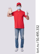Image of a happy young delivery man in red cap blank t-shirt uniform face mask gloves standing with empty brown craft paper packet isolated on light gray background studio, Service during pandemic coronavirus virus covid-19 concept. Стоковое фото, агентство Ingram Publishing / Фотобанк Лори