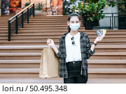 Portrait of young Asian woman in protection mask going down stairs of shopping mall  with shopping paper bag. She show credit card in her hand. Стоковое фото, агентство Ingram Publishing / Фотобанк Лори