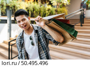 Portrait Handsome young man grinning as he holds a paper bag and shows his credit card on the stairs of a department store. New Normal lifestyle and shopping. Стоковое фото, агентство Ingram Publishing / Фотобанк Лори