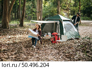 Pretty woman sitting on chair at front of camping tent and checking direction on paper map, Handsome boyfriend is pitching a tent behind her, They are happy to camping in forest on vacation . Стоковое фото, агентство Ingram Publishing / Фотобанк Лори