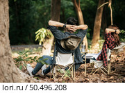 Rear view, Young Asian teenager couple have relax time with camping trip, They are sitting and hands on back of neck on chair in front of backpack camping tent in forest. Стоковое фото, агентство Ingram Publishing / Фотобанк Лори