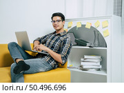 Young handsome man wearing eyeglasses with new normal, sitting on sofa and works from home on laptop computer while self-isolation and quarantine covid-19 coronavirus outbreak. Стоковое фото, агентство Ingram Publishing / Фотобанк Лори