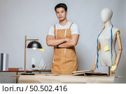 Portrait Young male fashion designer working with fabric and clothing sketches at the studio at home, full of tailoring tools, model and equipment on table. Стоковое фото, агентство Ingram Publishing / Фотобанк Лори