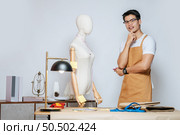 Portrait Young male fashion designer in glasses working with mannequin and clothing sketches for lady at the studio at home,  full of tailoring tools and equipment. Стоковое фото, агентство Ingram Publishing / Фотобанк Лори