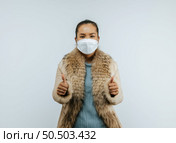 Portrait of Asian woman with two thumbs up sign gesture, putting on protective mask on face for protection against coronavirus, wearing a fur coat standing over background in studio with copy space. Стоковое фото, агентство Ingram Publishing / Фотобанк Лори