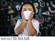 Portrait of a little girl wearing eyeglasses and facemask at school to avoid the spread of coronavirus, new normal with schoolchild,  blure blackboard in background. Стоковое фото, агентство Ingram Publishing / Фотобанк Лори