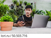 Happily gardener man use smartphone and laptop computer while online tutorial on potted plans in shop, vlogging or small business houseplant concept. Стоковое фото, агентство Ingram Publishing / Фотобанк Лори