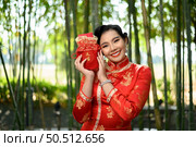Portrait pretty Asian woman in a Chinese cheongsam holding envelopes in Red amulet bag with Chinese alphabet means good fortune and happinese in bamboo forest background, Chinese New Year concept. Стоковое фото, агентство Ingram Publishing / Фотобанк Лори
