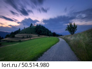 Rural road and chapel on the mountain peak at twilight. Gravel road, green meadows and trees, church on the hill and purple sky with blurred clouds at sunset in Slovenia. Nature. Summer night. Стоковое фото, агентство Ingram Publishing / Фотобанк Лори