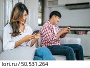 Selective focus, Young Asian Couple sitting on sofa in living room, They are enjoy with social media networks addicts surf the internet on mobile phones ignoring each other, family addicted smartphone concept. Стоковое фото, агентство Ingram Publishing / Фотобанк Лори