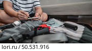 Asian man is preparing clothes in suitcases. He’s choosing clothes, travel documents, the itinerary for a solo trip, and check the checklist. Travel, holiday and vacation concept. Стоковое фото, агентство Ingram Publishing / Фотобанк Лори