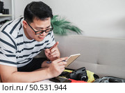 Asian man glasses is preparing clothes in suitcases. He’s choosing clothes, travel documents, the itinerary for a solo trip, and check the checklist. Travel, holiday and vacation concept. Стоковое фото, агентство Ingram Publishing / Фотобанк Лори