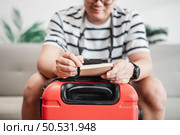 Asian man glasses is preparing clothes in suitcases. He’s choosing clothes, travel documents, the itinerary for a solo trip, and check the checklist. Travel, holiday and vacation concept. Стоковое фото, агентство Ingram Publishing / Фотобанк Лори