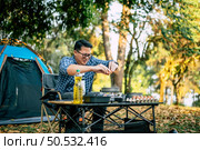 Broken egg falls into the frying pan at the campsite. Happy Asian traveler man cooking with tent background. Outdoor cooking, traveling, camping, lifestyle concept. Стоковое фото, агентство Ingram Publishing / Фотобанк Лори