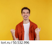 Portrait of happy joyful young man standing doing winner gesture clenching fists keeping isolated on yellow color wall background studio. Victory and success concept. Стоковое фото, агентство Ingram Publishing / Фотобанк Лори