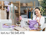Glad young Asian tailoress viewing dress detail sitting at sewing-machine. Стоковое фото, фотограф Яков Филимонов / Фотобанк Лори