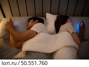 Young couple sleeping on the bed in the bedroom at night time with their backs to each other and playing on their phones, ignoring each other. Стоковое фото, агентство Ingram Publishing / Фотобанк Лори