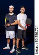 Full length portrait of two sporty young men in sportswear standing on indoor court with paddle rackets in hands after match. Стоковое фото, агентство Ingram Publishing / Фотобанк Лори